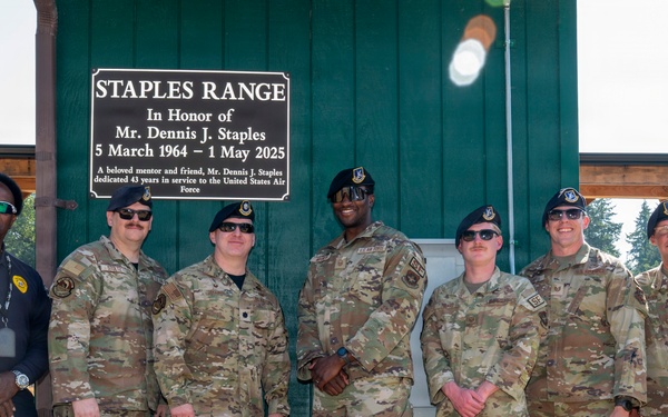 A group of seven individuals, including six military personnel in OCP (Operational Camouflage Pattern) uniforms and one civilian, pose in front of a dedication plaque at 'Staples Range,' honoring Mr. Dennis J. Staples (5 March 1964 – 1 May 2025). The plaque notes he dedicated 43 years in service to the United States Air Force. The setting appears to be an outdoor military shooting or training range.