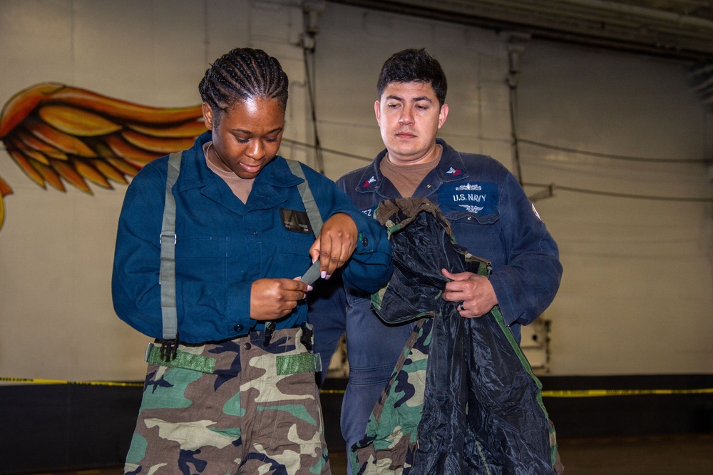 Two U.S. Navy personnel are examining equipment in what appears to be a hangar or indoor military facility. One sailor wearing camouflage pants and a blue coverall top is inspecting a strap or piece of gear, while the other, wearing a U.S. Navy-labeled blue coverall, holds what appears to be a harness or tactical vest. A painted mural featuring wings is visible on the wall in the background.