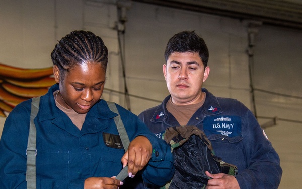Two U.S. Navy personnel are examining equipment in what appears to be a hangar or indoor military facility. One sailor wearing camouflage pants and a blue coverall top is inspecting a strap or piece of gear, while the other, wearing a U.S. Navy-labeled blue coverall, holds what appears to be a harness or tactical vest. A painted mural featuring wings is visible on the wall in the background.