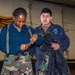 Two U.S. Navy personnel are examining equipment in what appears to be a hangar or indoor military facility. One sailor wearing camouflage pants and a blue coverall top is inspecting a strap or piece of gear, while the other, wearing a U.S. Navy-labeled blue coverall, holds what appears to be a harness or tactical vest. A painted mural featuring wings is visible on the wall in the background.