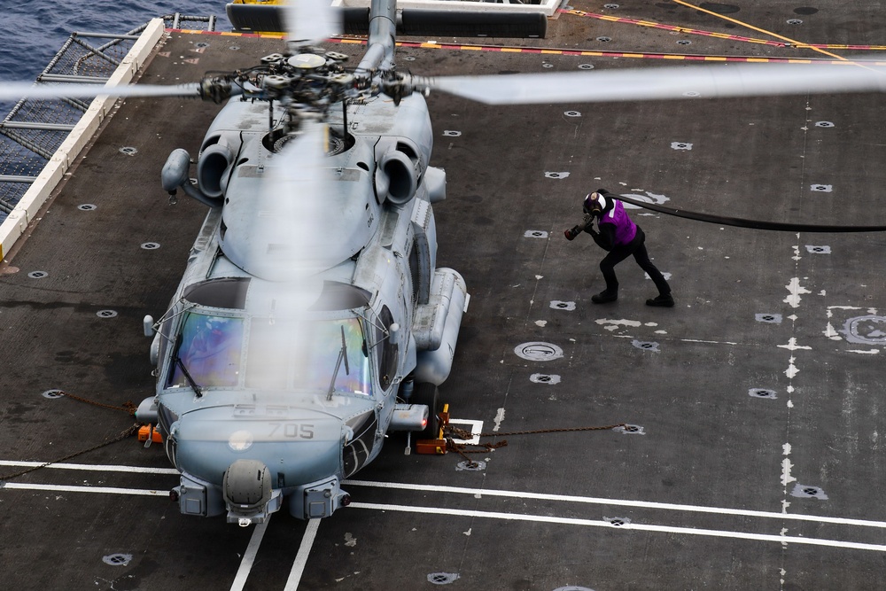 An aerial view of a military helicopter on a naval vessel's flight deck, with a crew member in a purple vest handling a fuel hose. The helicopter appears to be an MH-60 Seahawk variant, secured to the deck with tie-downs, and is undergoing a hot refueling or servicing operation. The image is taken from an elevated angle, likely from the ship's island or an overhead structure.