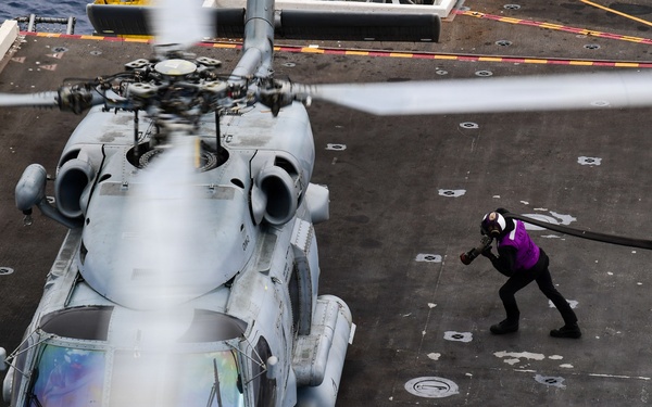 An aerial view of a military helicopter on a naval vessel's flight deck, with a crew member in a purple vest handling a fuel hose. The helicopter appears to be an MH-60 Seahawk variant, secured to the deck with tie-downs, and is undergoing a hot refueling or servicing operation. The image is taken from an elevated angle, likely from the ship's island or an overhead structure.