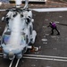An aerial view of a military helicopter on a naval vessel's flight deck, with a crew member in a purple vest handling a fuel hose. The helicopter appears to be an MH-60 Seahawk variant, secured to the deck with tie-downs, and is undergoing a hot refueling or servicing operation. The image is taken from an elevated angle, likely from the ship's island or an overhead structure.