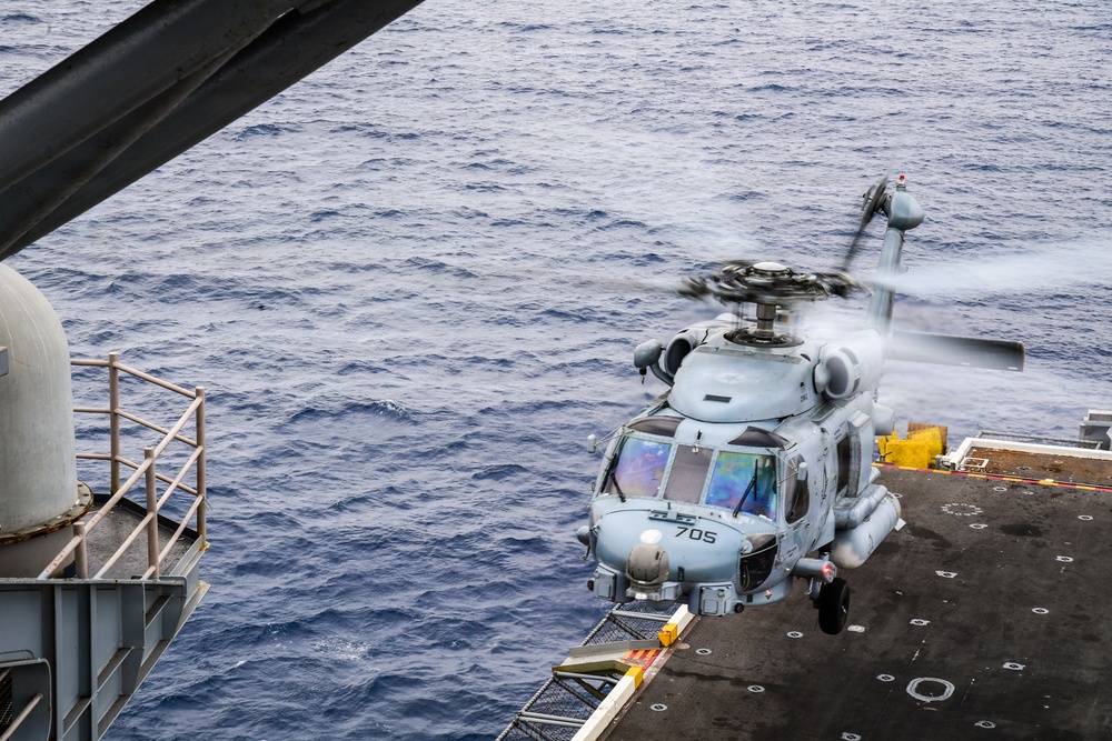 A military helicopter, marked with the number '705', is captured in the process of landing on or departing from a naval vessel's flight deck over open ocean. The helicopter appears to be a Sikorsky MH-60 Sea Hawk variant, painted in light gray naval livery. The rotor blades are in motion, and the ocean is visible in the background below the ship's deck level.