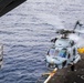 A military helicopter, marked with the number '705', is captured in the process of landing on or departing from a naval vessel's flight deck over open ocean. The helicopter appears to be a Sikorsky MH-60 Sea Hawk variant, painted in light gray naval livery. The rotor blades are in motion, and the ocean is visible in the background below the ship's deck level.