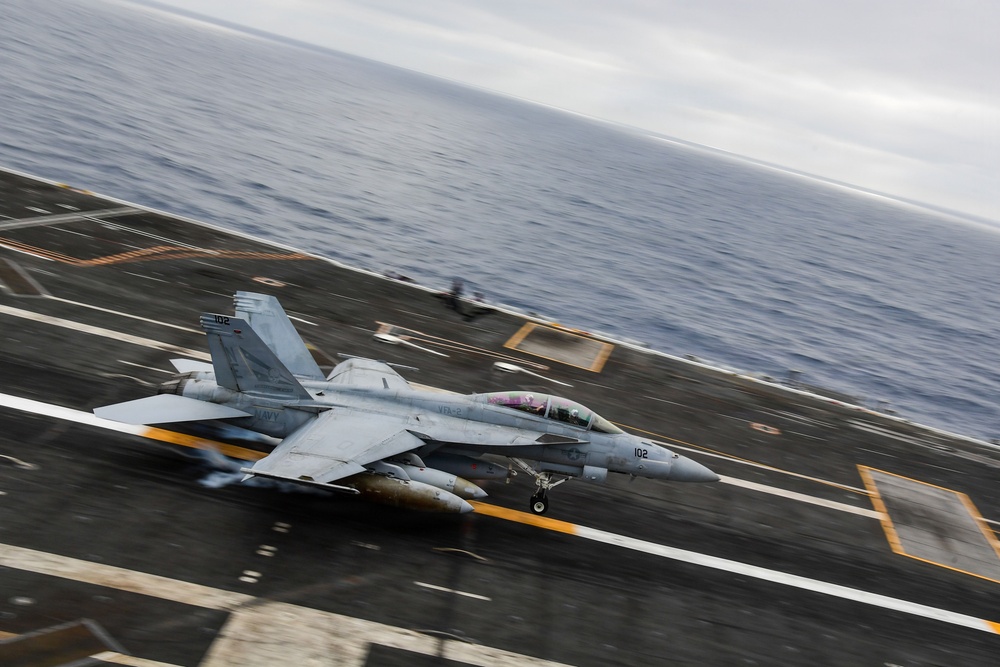 A U.S. Navy F/A-18 Super Hornet (bureau number 102) is captured launching or landing on an aircraft carrier flight deck over open ocean. The image is taken from an elevated angle showing the full flight deck with motion blur indicating high speed. A second aircraft is faintly visible in the background further down the deck.