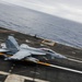A U.S. Navy F/A-18 Super Hornet (bureau number 102) is captured launching or landing on an aircraft carrier flight deck over open ocean. The image is taken from an elevated angle showing the full flight deck with motion blur indicating high speed. A second aircraft is faintly visible in the background further down the deck.