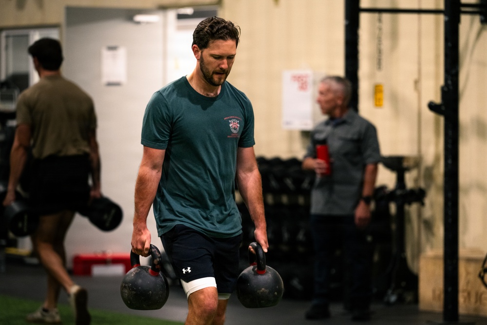 A man in a teal t-shirt and black Under Armour shorts carries two heavy kettlebells in a gym setting, performing what appears to be a farmer's carry exercise. He has a white bandage wrap on one knee and appears to be mid-workout with sweat-dampened hair. Several other individuals are visible in the blurred background of what appears to be a functional fitness or CrossFit-style gym.