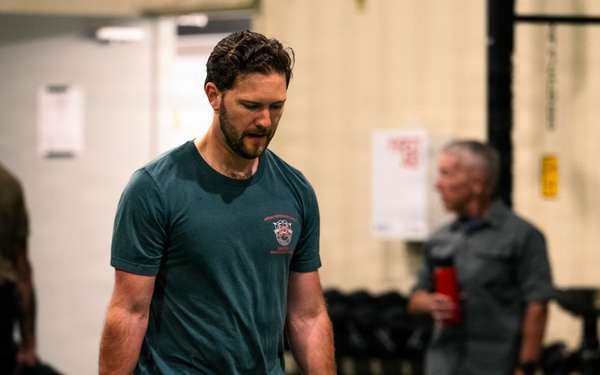 A man in a teal t-shirt and black Under Armour shorts carries two heavy kettlebells in a gym setting, performing what appears to be a farmer's carry exercise. He has a white bandage wrap on one knee and appears to be mid-workout with sweat-dampened hair. Several other individuals are visible in the blurred background of what appears to be a functional fitness or CrossFit-style gym.