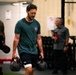 A man in a teal t-shirt and black Under Armour shorts carries two heavy kettlebells in a gym setting, performing what appears to be a farmer's carry exercise. He has a white bandage wrap on one knee and appears to be mid-workout with sweat-dampened hair. Several other individuals are visible in the blurred background of what appears to be a functional fitness or CrossFit-style gym.