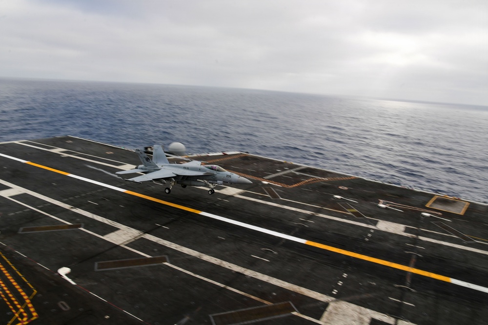 A carrier-based jet aircraft is captured mid-landing or takeoff roll on the flight deck of a naval aircraft carrier at sea. The aircraft, marked with the number '206', is positioned on the centerline of the angled flight deck with the open ocean visible in the background. Overcast sky conditions and motion blur suggest dynamic operational activity.