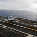 A carrier-based jet aircraft is captured mid-landing or takeoff roll on the flight deck of a naval aircraft carrier at sea. The aircraft, marked with the number '206', is positioned on the centerline of the angled flight deck with the open ocean visible in the background. Overcast sky conditions and motion blur suggest dynamic operational activity.