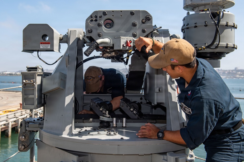 Two U.S. Navy personnel are performing maintenance or inspection work on what appears to be a Close-In Weapon System (CIWS) or similar naval gun mount aboard a warship docked at a pier. One sailor is leaning inside the open housing of the weapon system while the other operates a manual control handle on the exterior. The setting appears to be a naval base with calm water and a shoreline visible in the background.