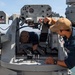 Two U.S. Navy personnel are performing maintenance or inspection work on what appears to be a Close-In Weapon System (CIWS) or similar naval gun mount aboard a warship docked at a pier. One sailor is leaning inside the open housing of the weapon system while the other operates a manual control handle on the exterior. The setting appears to be a naval base with calm water and a shoreline visible in the background.