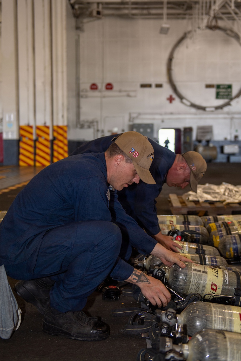 Two personnel in navy blue coveralls and tan caps are crouched down inspecting or servicing a row of Self-Contained Breathing Apparatus (SCBA) air cylinders, identifiable as Scott brand units. The setting appears to be a hangar bay or large industrial space aboard a naval vessel, indicated by the USS cap insignia and characteristic ship interior markings. The activity suggests routine maintenance, inspection, or inventory of firefighting/damage control equipment.