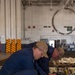 Two personnel in navy blue coveralls and tan caps are crouched down inspecting or servicing a row of Self-Contained Breathing Apparatus (SCBA) air cylinders, identifiable as Scott brand units. The setting appears to be a hangar bay or large industrial space aboard a naval vessel, indicated by the USS cap insignia and characteristic ship interior markings. The activity suggests routine maintenance, inspection, or inventory of firefighting/damage control equipment.