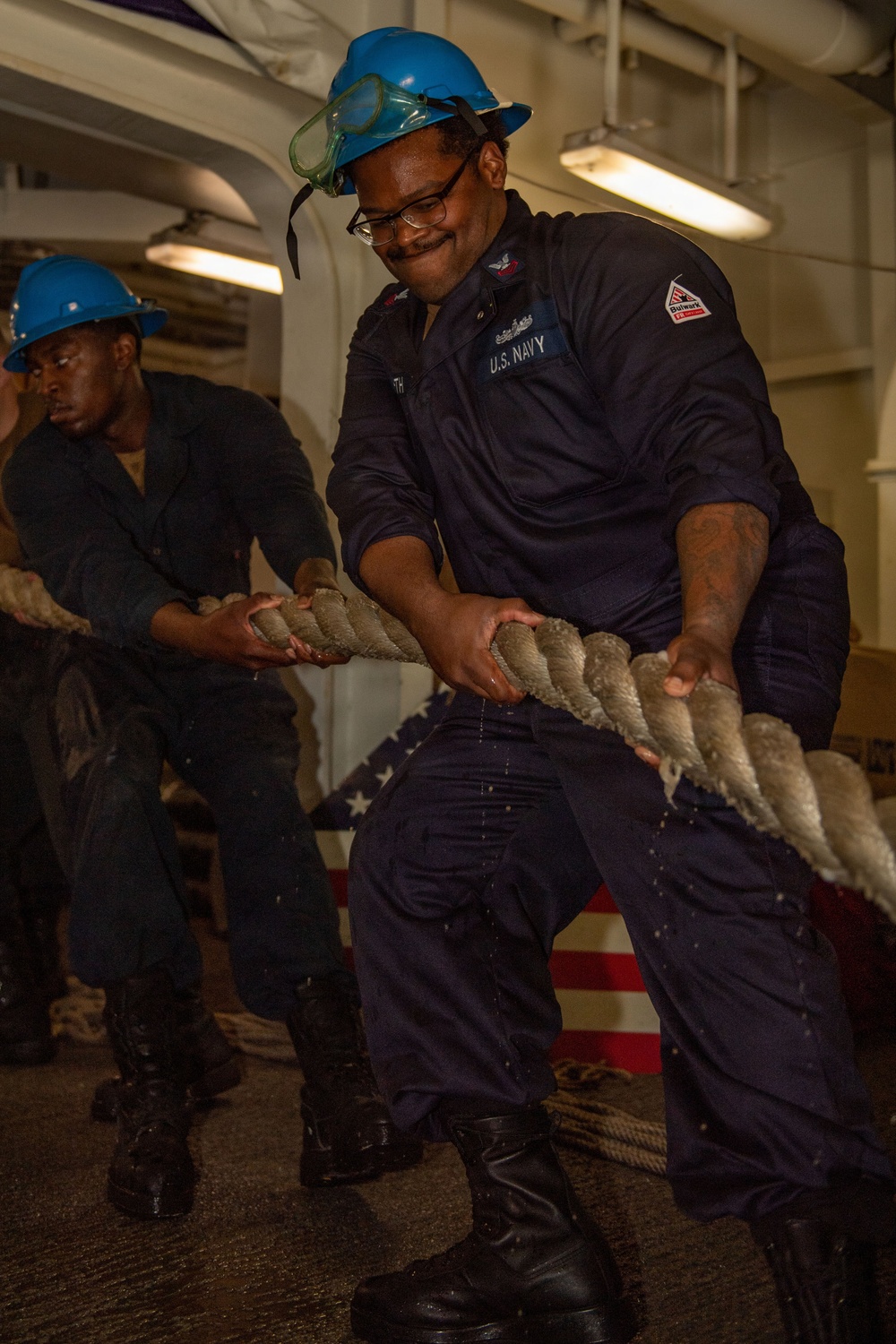 Two U.S. Navy sailors wearing blue hard hats and navy coveralls are pulling a large thick rope inside what appears to be a naval vessel. The foreground sailor is wearing safety goggles pushed up on his helmet and glasses, and is visibly exerting effort with a slight smile. An American flag is partially visible in the background.