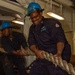 Two U.S. Navy sailors wearing blue hard hats and navy coveralls are pulling a large thick rope inside what appears to be a naval vessel. The foreground sailor is wearing safety goggles pushed up on his helmet and glasses, and is visibly exerting effort with a slight smile. An American flag is partially visible in the background.