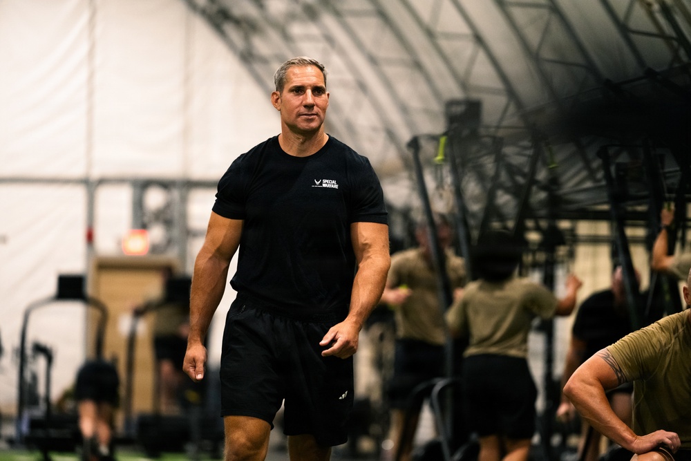 A physically fit middle-aged man wearing a black 'Special Warfare' t-shirt and black shorts walks through what appears to be a military or tactical fitness facility. He appears to be an instructor or supervisor overseeing a training session. In the background, multiple individuals in olive/tan military-style clothing are engaged in various physical training exercises.