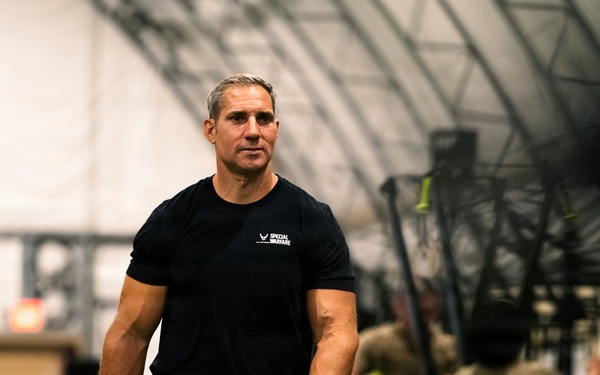 A physically fit middle-aged man wearing a black 'Special Warfare' t-shirt and black shorts walks through what appears to be a military or tactical fitness facility. He appears to be an instructor or supervisor overseeing a training session. In the background, multiple individuals in olive/tan military-style clothing are engaged in various physical training exercises.