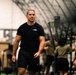 A physically fit middle-aged man wearing a black 'Special Warfare' t-shirt and black shorts walks through what appears to be a military or tactical fitness facility. He appears to be an instructor or supervisor overseeing a training session. In the background, multiple individuals in olive/tan military-style clothing are engaged in various physical training exercises.