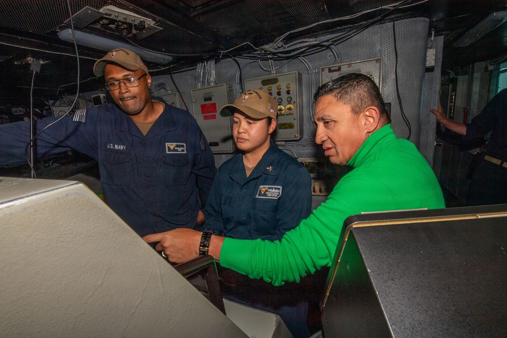 Three individuals are gathered around a control console aboard what appears to be a U.S. Navy vessel. Two personnel are wearing U.S. Navy uniforms with tan caps, while a third person in a green jacket appears to be a civilian or visitor. They are engaged in reviewing or operating equipment at the console.