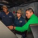 Three individuals are gathered around a control console aboard what appears to be a U.S. Navy vessel. Two personnel are wearing U.S. Navy uniforms with tan caps, while a third person in a green jacket appears to be a civilian or visitor. They are engaged in reviewing or operating equipment at the console.