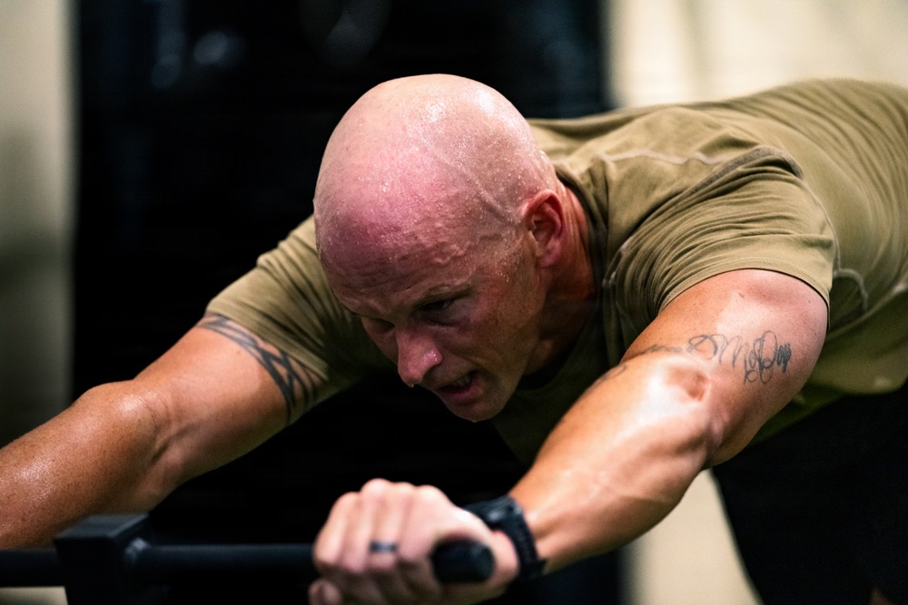 A muscular, bald man is intensely exercising on gym equipment, visibly sweating heavily. He is leaning forward with both hands gripping handles, suggesting high-intensity cardio or strength training. His flushed skin and sweat indicate significant physical exertion.