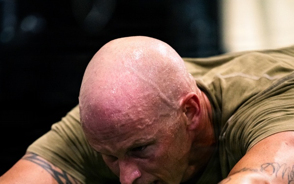 A muscular, bald man is intensely exercising on gym equipment, visibly sweating heavily. He is leaning forward with both hands gripping handles, suggesting high-intensity cardio or strength training. His flushed skin and sweat indicate significant physical exertion.
