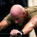 A muscular, bald man is intensely exercising on gym equipment, visibly sweating heavily. He is leaning forward with both hands gripping handles, suggesting high-intensity cardio or strength training. His flushed skin and sweat indicate significant physical exertion.