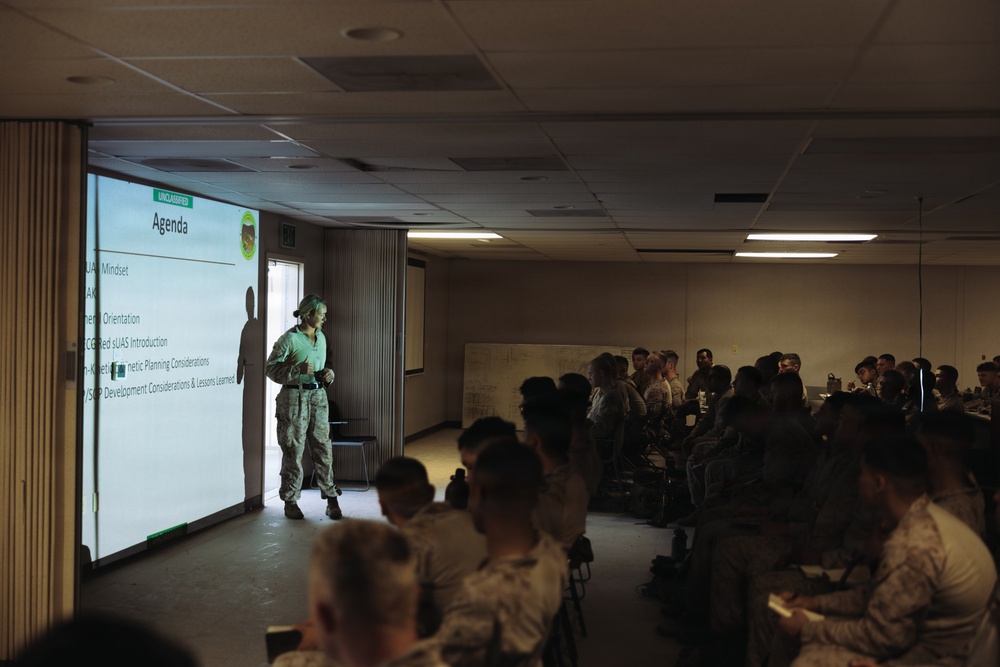 A military briefing or training session is underway in a dimly lit conference room. A presenter in military camouflage uniform stands near a projected slide titled 'Agenda' marked 'UNCLASSIFIED', addressing a room full of uniformed military personnel seated at tables. The agenda slide lists several topics including UAS-related subjects and planning considerations.