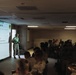 A military briefing or training session is underway in a dimly lit conference room. A presenter in military camouflage uniform stands near a projected slide titled 'Agenda' marked 'UNCLASSIFIED', addressing a room full of uniformed military personnel seated at tables. The agenda slide lists several topics including UAS-related subjects and planning considerations.