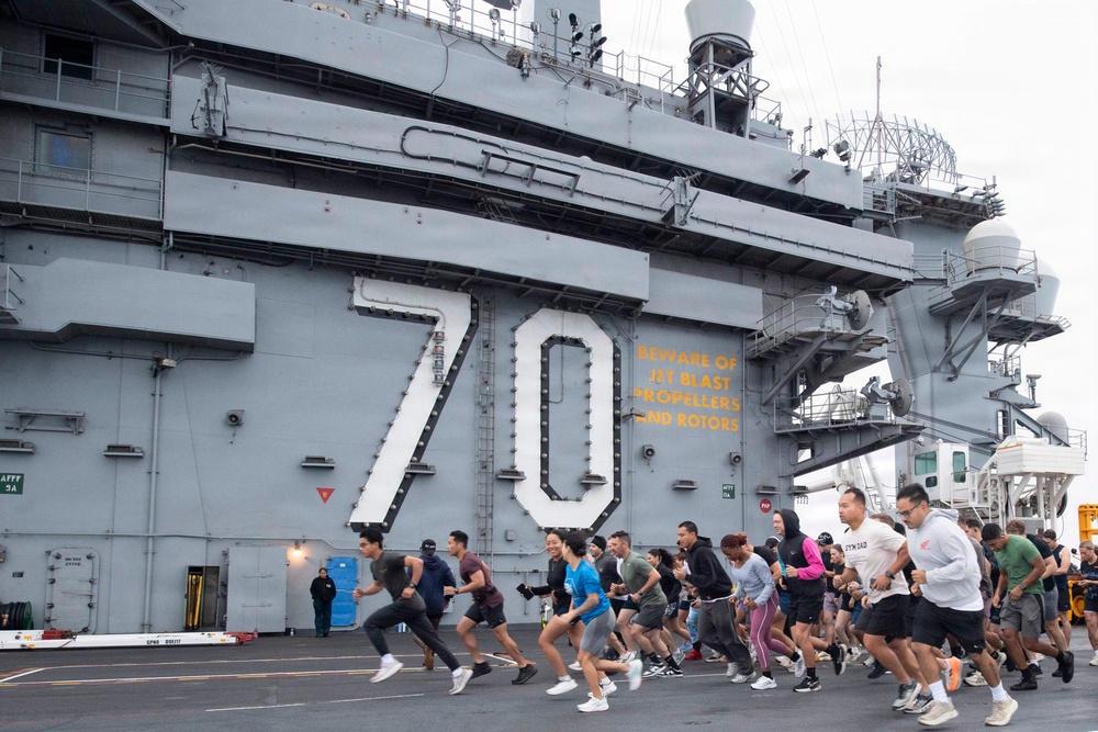 A large group of people are running on the flight deck of a U.S. Navy aircraft carrier bearing hull number 70, identified as the USS Carl Vinson (CVN-70). The runners appear to be participating in an organized fitness run or physical training event on the ship's deck. A warning sign reading 'BEWARE OF JET BLAST PROPELLERS AND ROTORS' is visible on the island superstructure.