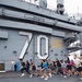 A large group of people are running on the flight deck of a U.S. Navy aircraft carrier bearing hull number 70, identified as the USS Carl Vinson (CVN-70). The runners appear to be participating in an organized fitness run or physical training event on the ship's deck. A warning sign reading 'BEWARE OF JET BLAST PROPELLERS AND ROTORS' is visible on the island superstructure.
