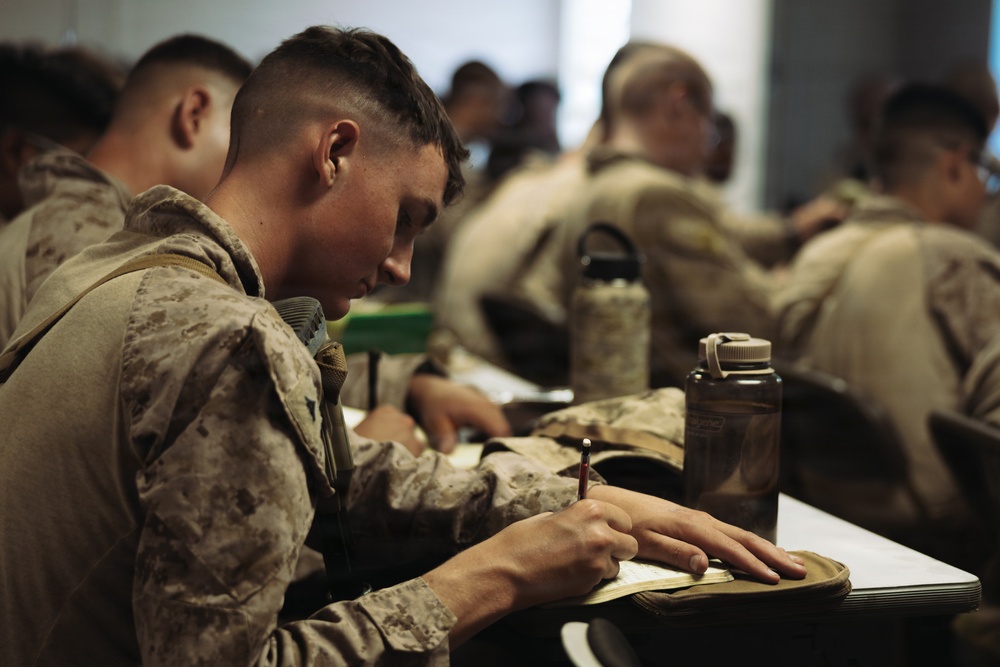 A group of military personnel in camouflage uniforms are seated at tables in what appears to be a classroom or briefing room setting. The foreground subject, a young male service member, is actively writing in a notebook with a pen. The image captures a training or instructional session, likely within a military educational environment.
