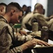 A group of military personnel in camouflage uniforms are seated at tables in what appears to be a classroom or briefing room setting. The foreground subject, a young male service member, is actively writing in a notebook with a pen. The image captures a training or instructional session, likely within a military educational environment.