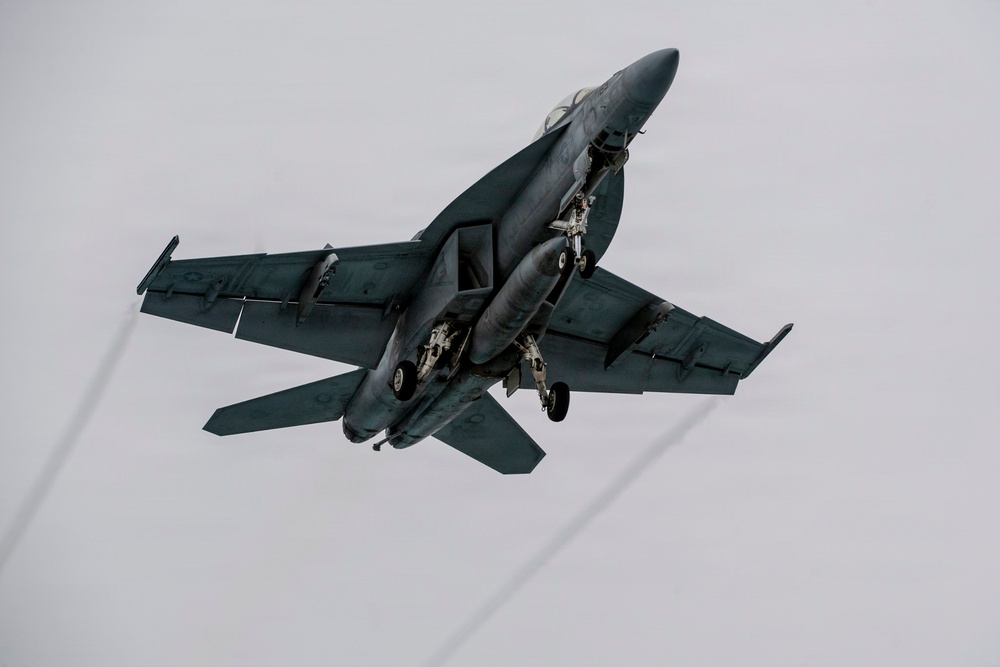 A military jet fighter, consistent with an F/A-18 Hornet or Super Hornet, is photographed from below at a steep banking angle against an overcast gray sky. The aircraft has its landing gear deployed, suggesting it is either taking off or preparing to land. The dark gray naval paint scheme and twin-engine configuration are clearly visible.