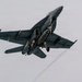 A military jet fighter, consistent with an F/A-18 Hornet or Super Hornet, is photographed from below at a steep banking angle against an overcast gray sky. The aircraft has its landing gear deployed, suggesting it is either taking off or preparing to land. The dark gray naval paint scheme and twin-engine configuration are clearly visible.