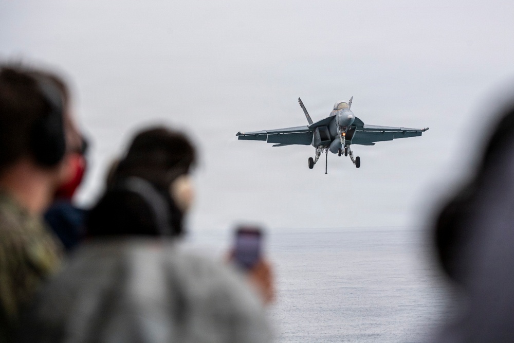 A military jet aircraft, consistent with an F/A-18 Hornet or Super Hornet, is captured on approach with landing gear deployed, flying low over open water. Several observers in the foreground, some wearing hearing protection, watch and photograph the aircraft. The scene appears to be from the deck or edge of a naval vessel.