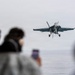 A military jet aircraft, consistent with an F/A-18 Hornet or Super Hornet, is captured on approach with landing gear deployed, flying low over open water. Several observers in the foreground, some wearing hearing protection, watch and photograph the aircraft. The scene appears to be from the deck or edge of a naval vessel.