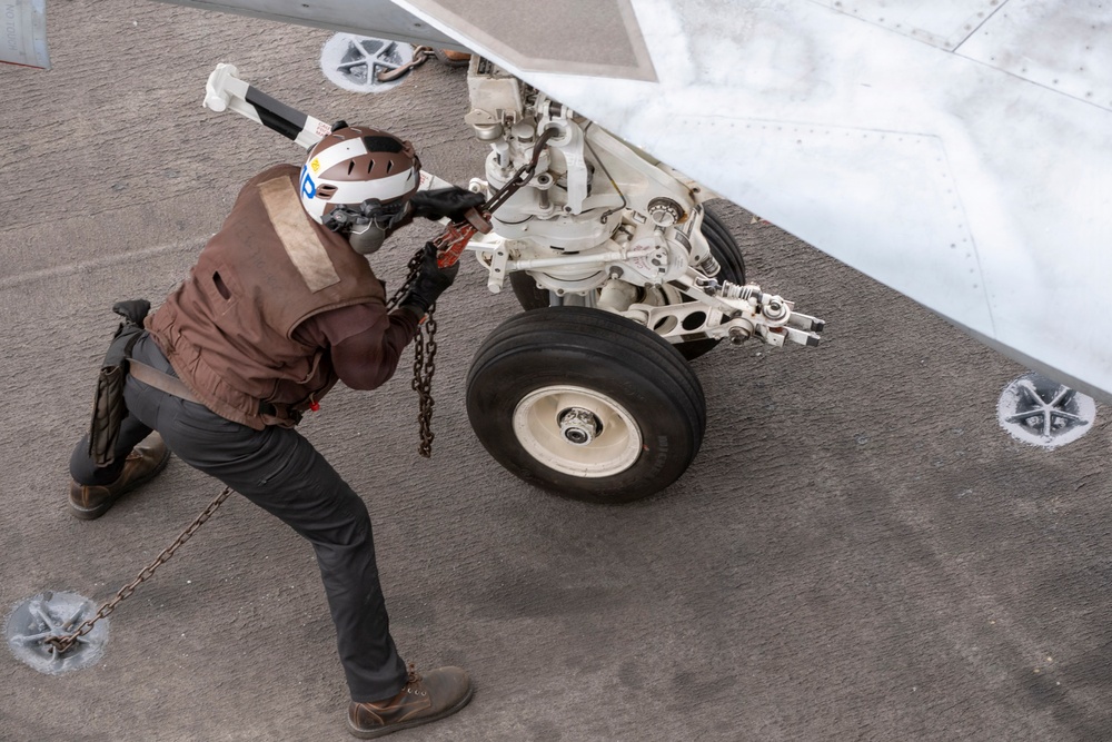 A naval deck crew member wearing a brown float coat and white helmet is working on the nose landing gear of a military aircraft on what appears to be an aircraft carrier flight deck. The technician is handling chains and appears to be attaching or detaching tie-down chains from the aircraft's landing gear assembly. The textured non-slip surface of the carrier deck and tie-down anchor points are clearly visible.