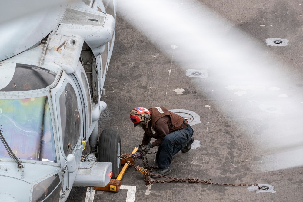 A deck crew member wearing a red helmet and brown jersey is crouching beside a military helicopter on what appears to be a naval vessel's flight deck. The worker is handling wheel chocks and tie-down chains securing the helicopter's landing gear to the deck. The image is taken from an elevated angle, partially obscured by what appears to be a rotor blade in the foreground.