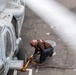 A deck crew member wearing a red helmet and brown jersey is crouching beside a military helicopter on what appears to be a naval vessel's flight deck. The worker is handling wheel chocks and tie-down chains securing the helicopter's landing gear to the deck. The image is taken from an elevated angle, partially obscured by what appears to be a rotor blade in the foreground.