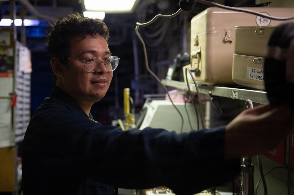 A young person wearing round wire-framed glasses and a dark navy uniform is working with equipment in what appears to be a military or industrial workspace. They are reaching toward stacked beige/cream-colored equipment cases or electronic units mounted on a rack. The environment is dimly lit with industrial lighting overhead.