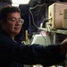 A young person wearing round wire-framed glasses and a dark navy uniform is working with equipment in what appears to be a military or industrial workspace. They are reaching toward stacked beige/cream-colored equipment cases or electronic units mounted on a rack. The environment is dimly lit with industrial lighting overhead.