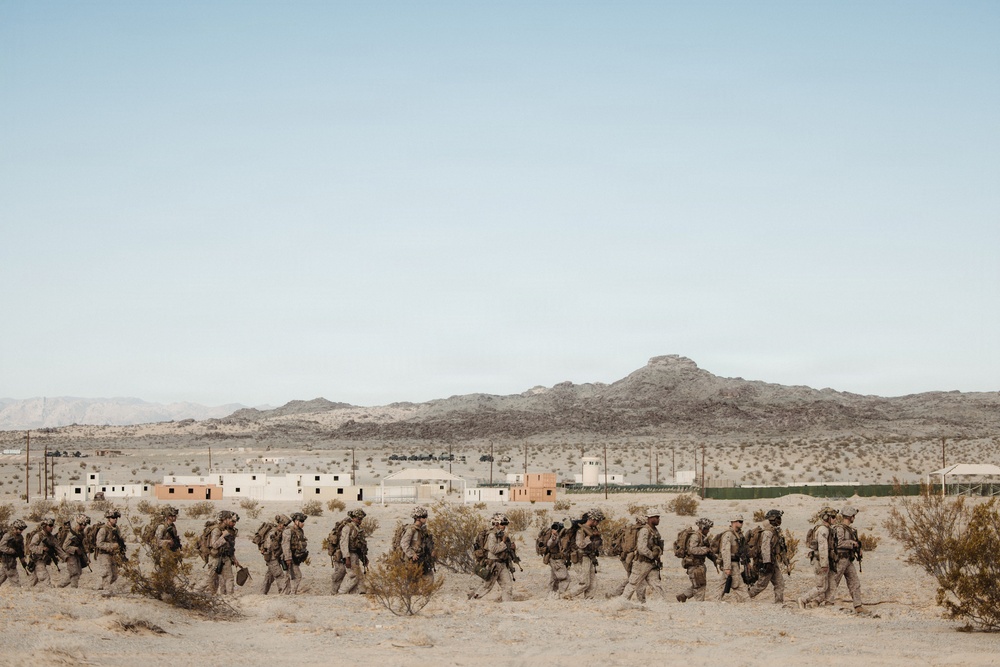 A column of military personnel in desert camouflage uniforms and full combat gear marches in single file through an arid desert landscape. In the background, a cluster of low-rise buildings consistent with a military training compound or mock village is visible, set against rocky desert mountains. The scene strongly suggests a military training exercise in a desert environment.