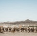 A column of military personnel in desert camouflage uniforms and full combat gear marches in single file through an arid desert landscape. In the background, a cluster of low-rise buildings consistent with a military training compound or mock village is visible, set against rocky desert mountains. The scene strongly suggests a military training exercise in a desert environment.