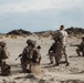 A group of military personnel in desert camouflage uniforms are conducting what appears to be a training exercise in an arid desert environment. One standing individual, without a helmet, appears to be an instructor or leader giving directional hand signals to the kneeling soldiers. The terrain is flat, sandy, and sparse with rocky outcroppings visible in the background.