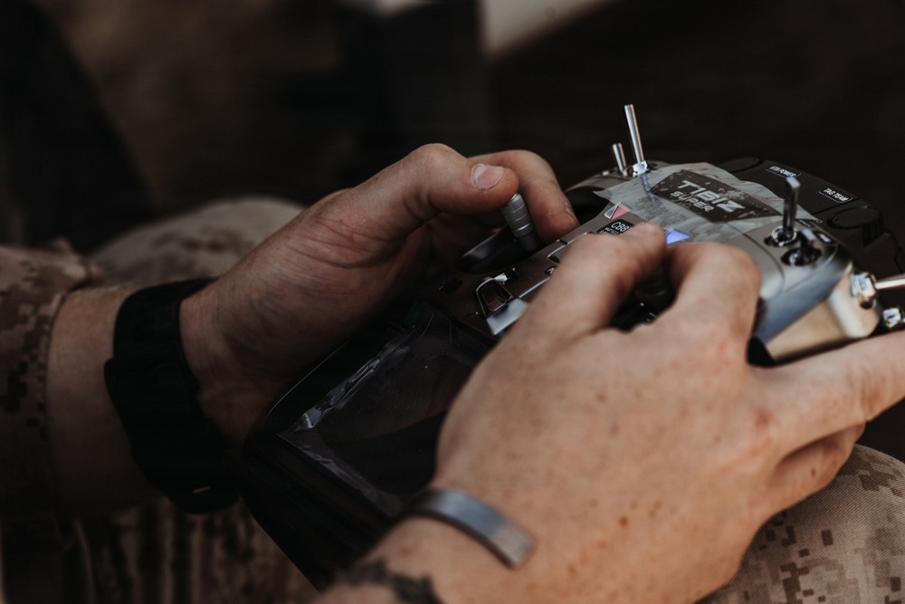A person wearing military camouflage clothing is operating a radio transmitter/remote control unit, identifiable as a Radiomaster or similar brand TX16S SUPER model. The individual has a gray/blue wristband and is manipulating the control sticks and switches on the device. The close-up shot focuses on the hands and controller, with the background blurred.