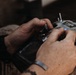 A person wearing military camouflage clothing is operating a radio transmitter/remote control unit, identifiable as a Radiomaster or similar brand TX16S SUPER model. The individual has a gray/blue wristband and is manipulating the control sticks and switches on the device. The close-up shot focuses on the hands and controller, with the background blurred.